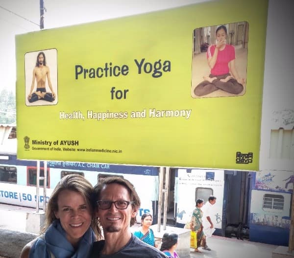 William and Elizabeth in India at a train station with 'Practice Yoga for Health, Happiness and Harmony' sign